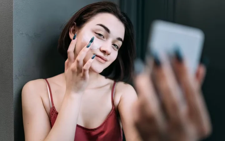 A woman applying moisturiser to her face while holding a phone