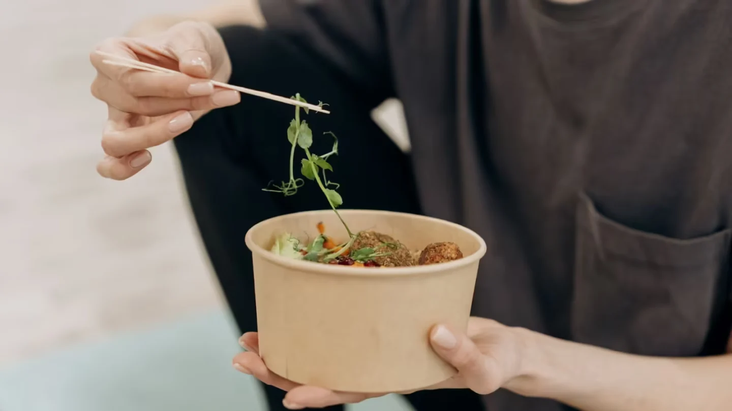 Woman holding a paper bowl filled with a healthy vegetarian meal in an indoor setting, promoting wellness.