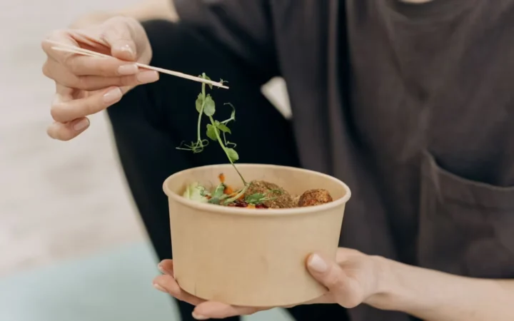 A woman holding a paper bowl filled with a healthy vegetarian meal in an indoor setting, promoting wellness.