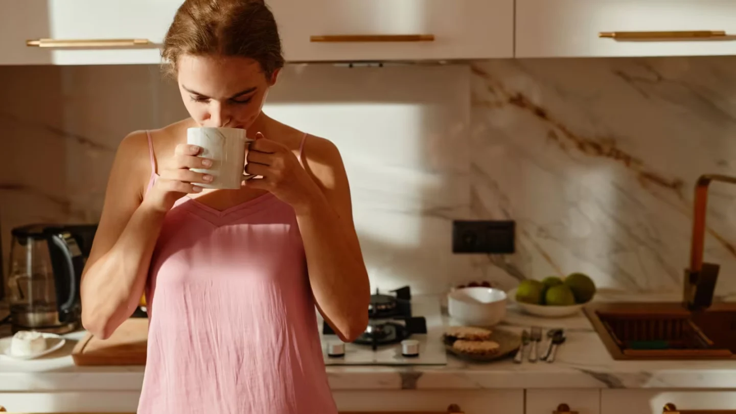 Woman enjoys a hot beverage in a sunlit kitchen, creating a tranquil morning scene.