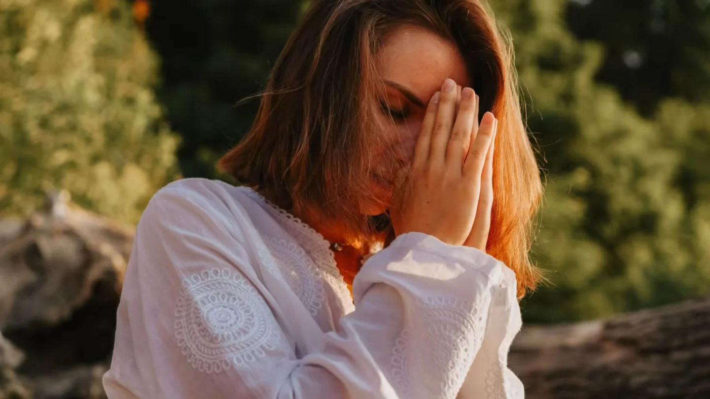 Woman meditating outdoors, eyes closed in peaceful prayer pose.