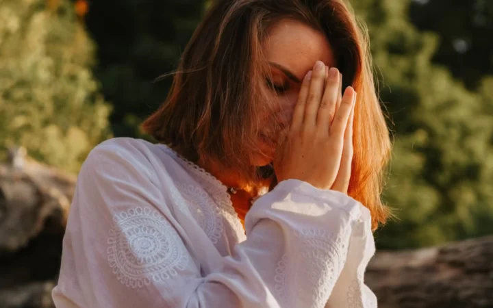Woman meditating outdoors, eyes closed in peaceful prayer pose.