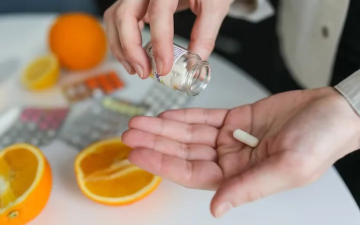 Hands tipping a supplement tablet from a bottle beside sliced oranges