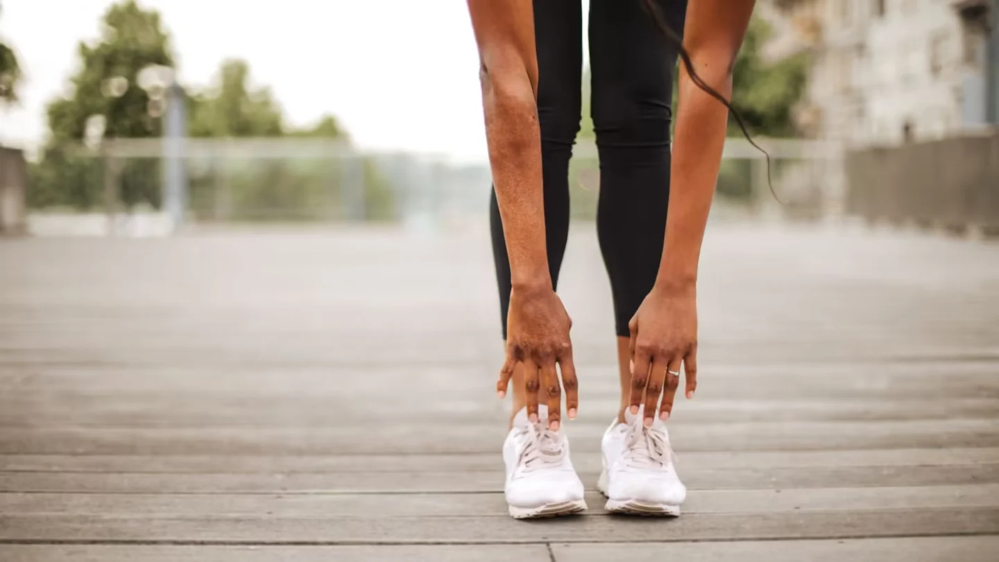 From below crop slender female athlete in sportswear and white sneakers doing standing forward bend exercise for stretching