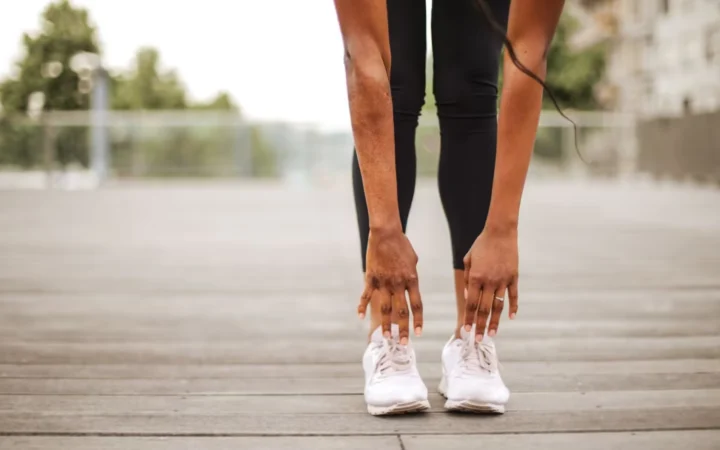 From below crop slender female athlete in sportswear and white sneakers doing standing forward bend exercise for stretching body on wooden floor of street sports ground against blurred urban environment in daytime
