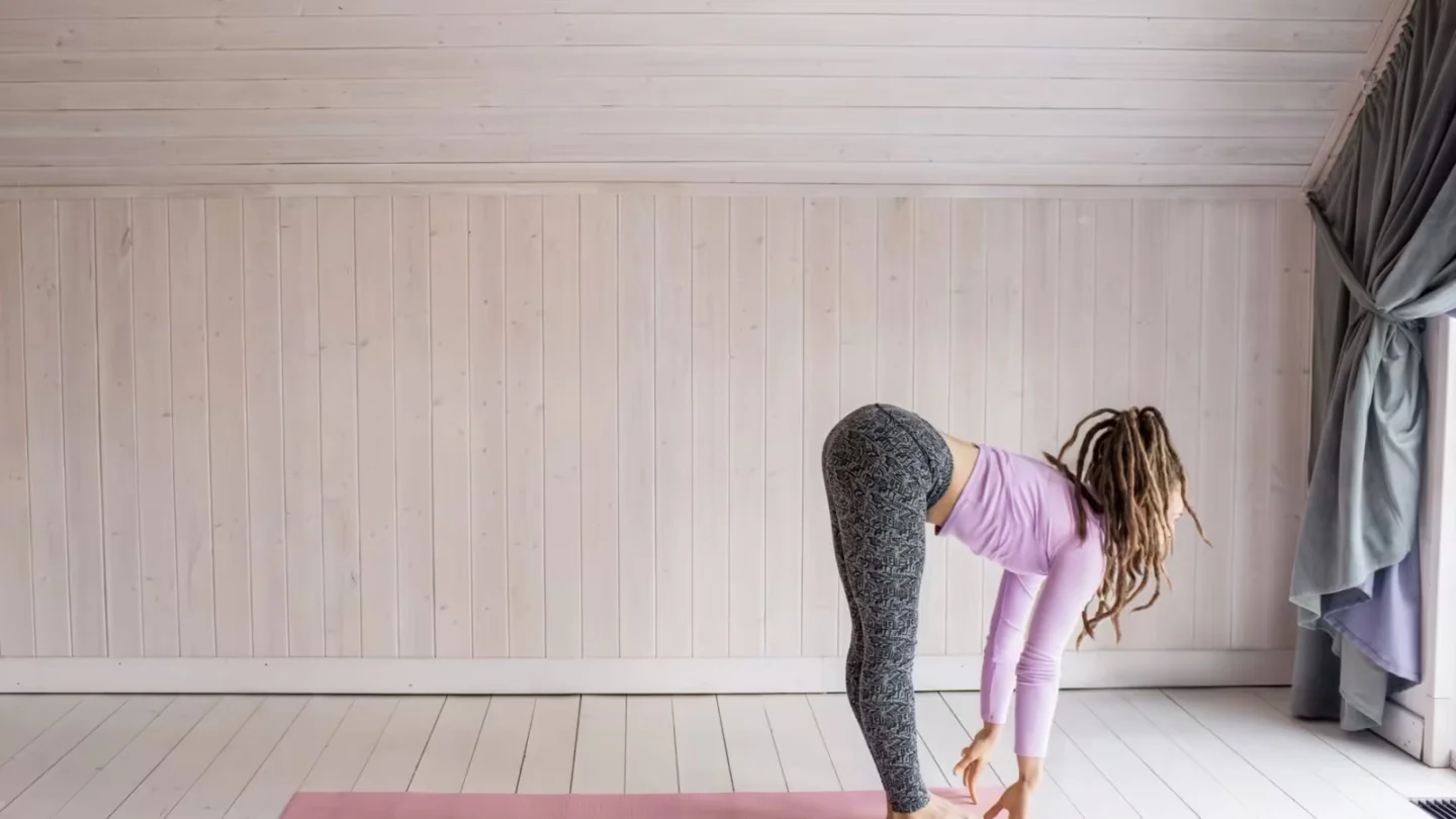 Woman with dreadlocks performs a forward bend yoga pose on a pink mat indoors.