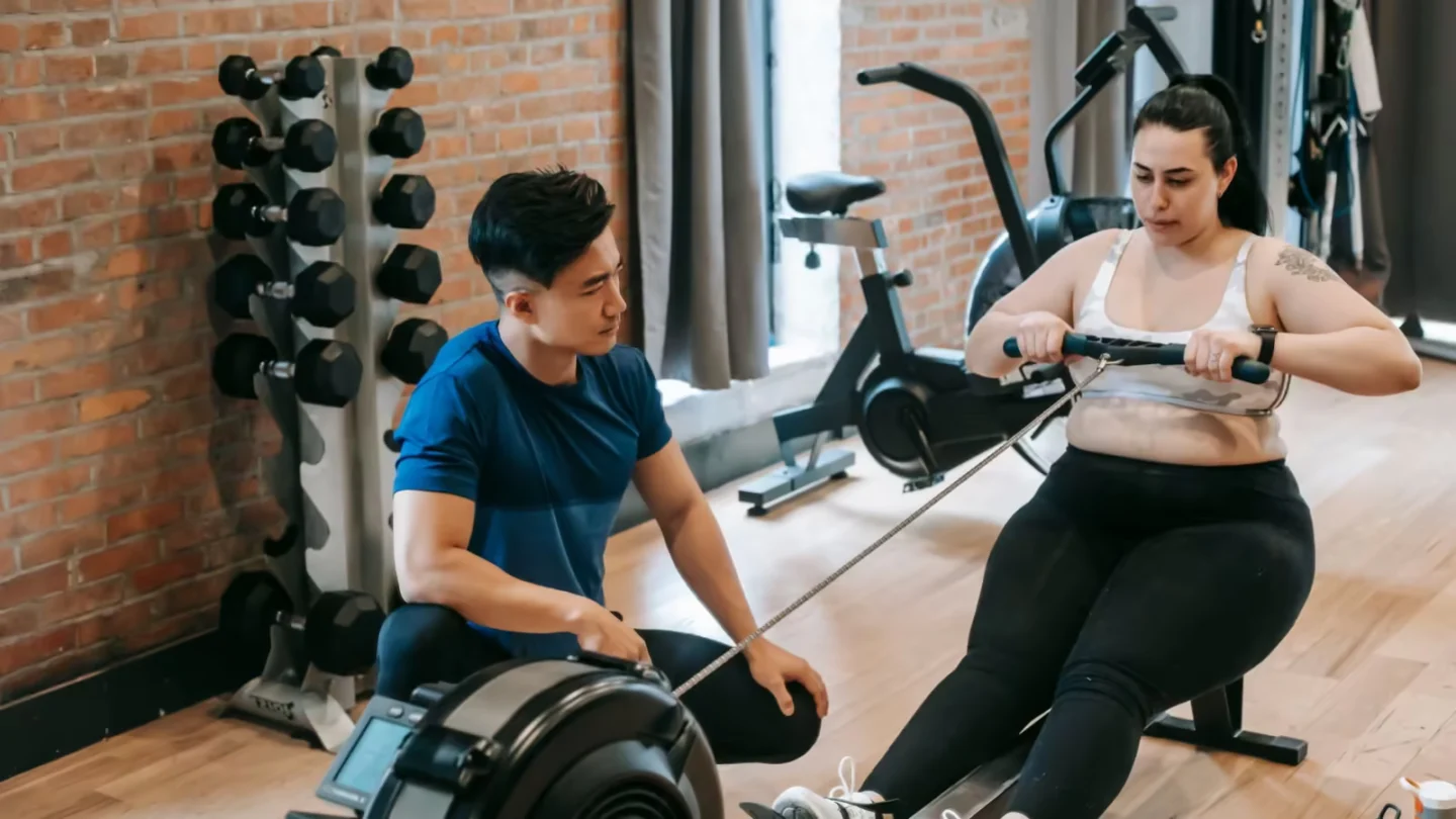 From above of professional ethnic constructor helping young plus size woman working out with rowing machine in sport club