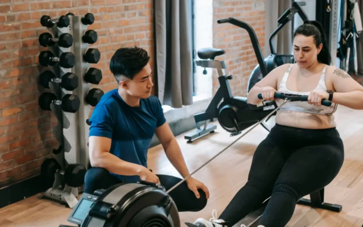 From above of professional ethnic constructor helping young plus size woman working out with rowing machine in sport club