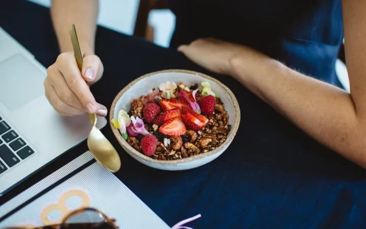 A bowl of granola with strawberries and blueberries beside a laptop