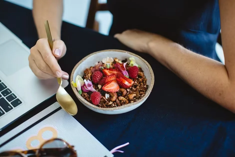 A bowl of granola with strawberries and blueberries beside a laptop