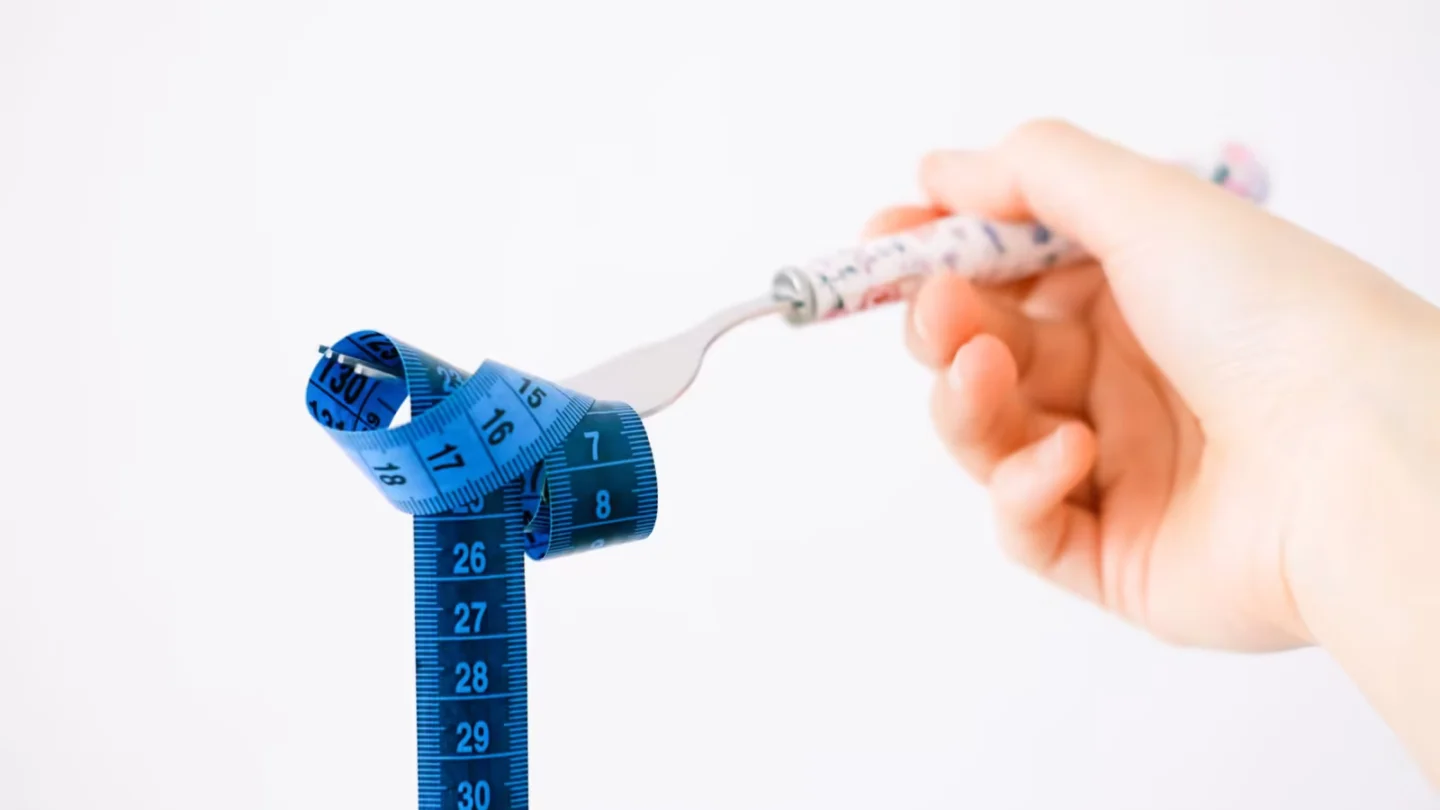 Hand holding a fork wrapped with a blue measuring tape suggesting diet or weight management.