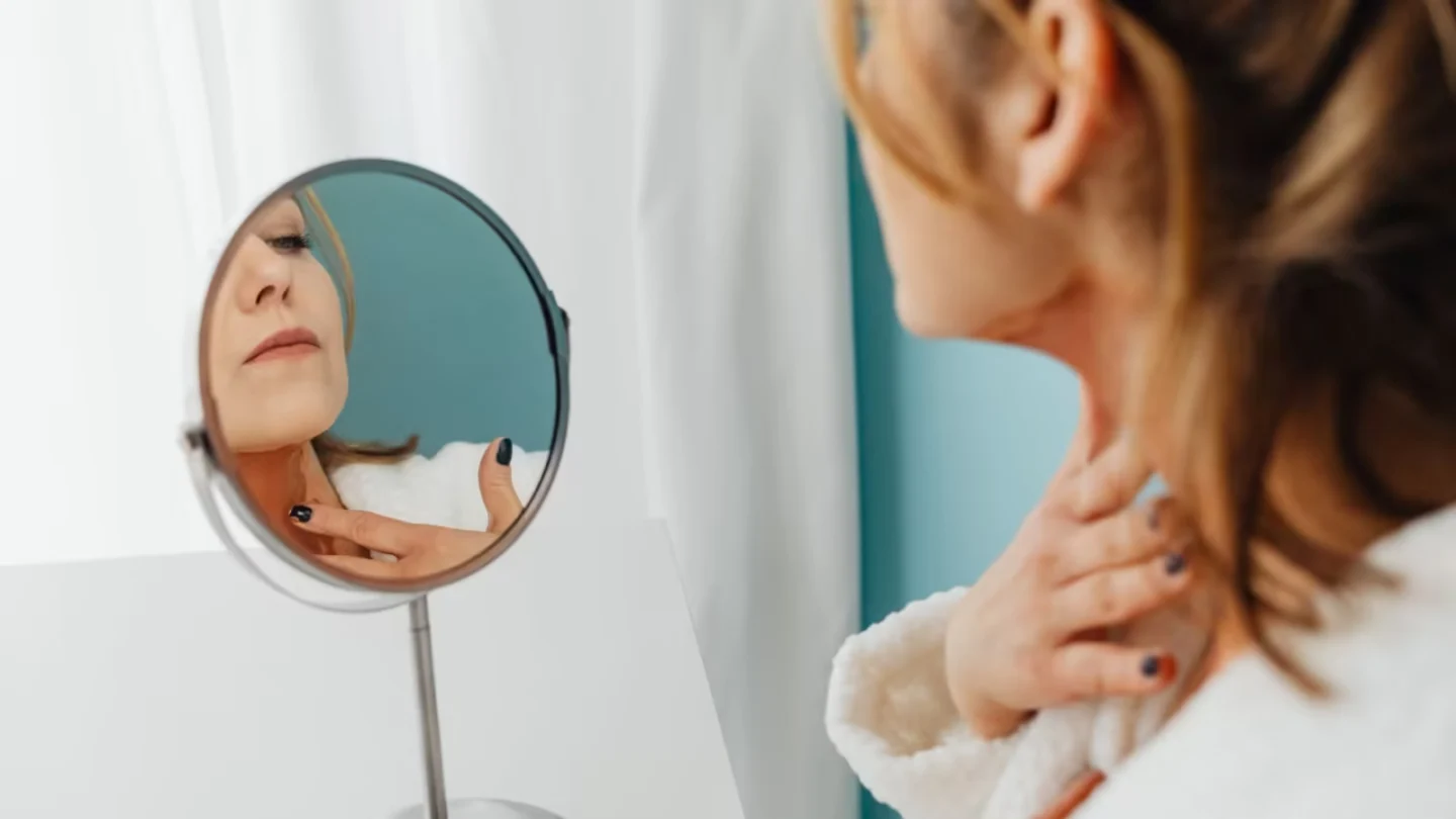 Woman in a bathrobe applying skincare while looking in the mirror.