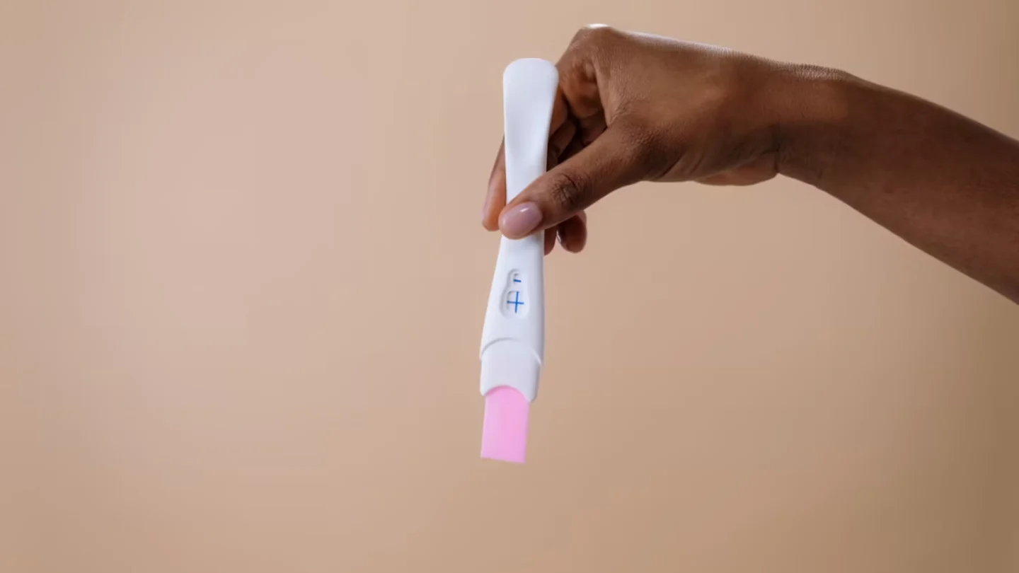 Woman's hand holding a positive pregnancy test on a neutral background.