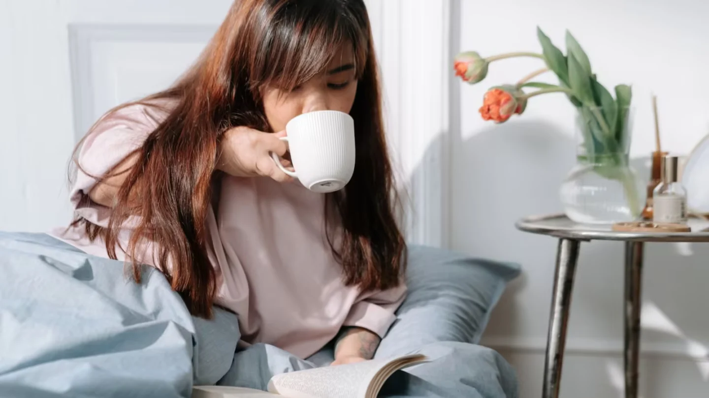Asian woman enjoying her morning with coffee and a book in a cozy bedroom setting.