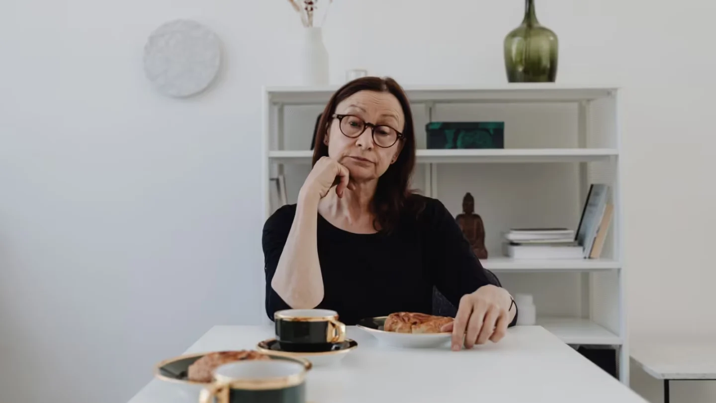Elderly woman sits pensively at a white table with coffee and pastries.