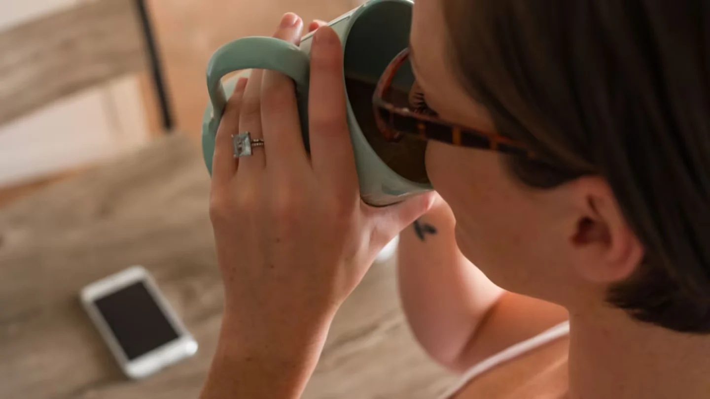 Woman with glasses drinks coffee from a mug indoors, smartphone on table.