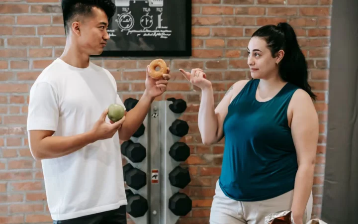 Side view of Asian personal instructor with apple and doughnut offering healthy food to plus size woman while standing in gym