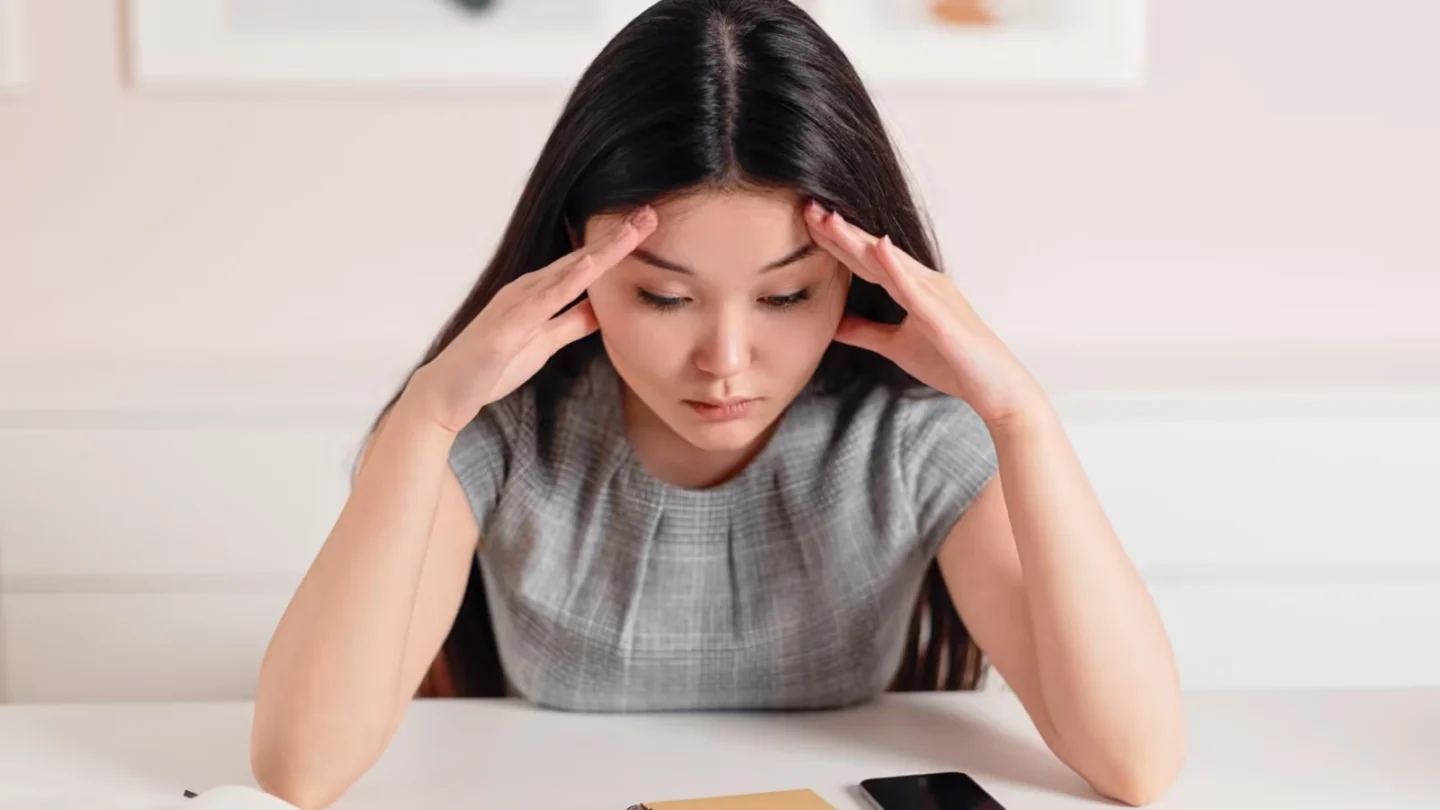 Asian businesswoman with a headache at her desk, displaying stress and anxiety.