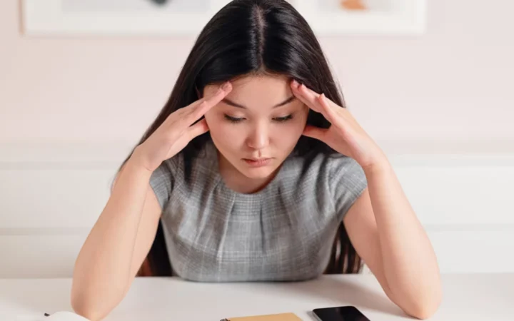 Asian businesswoman with a headache at her desk, displaying stress and anxiety.