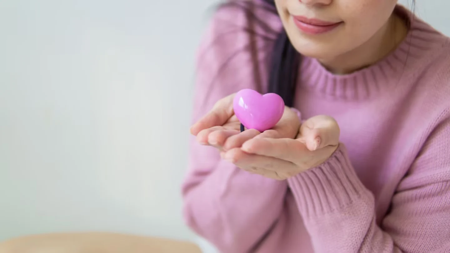Woman in a pink sweater holding a pink heart-shaped toy, symbolizing love and kindness.