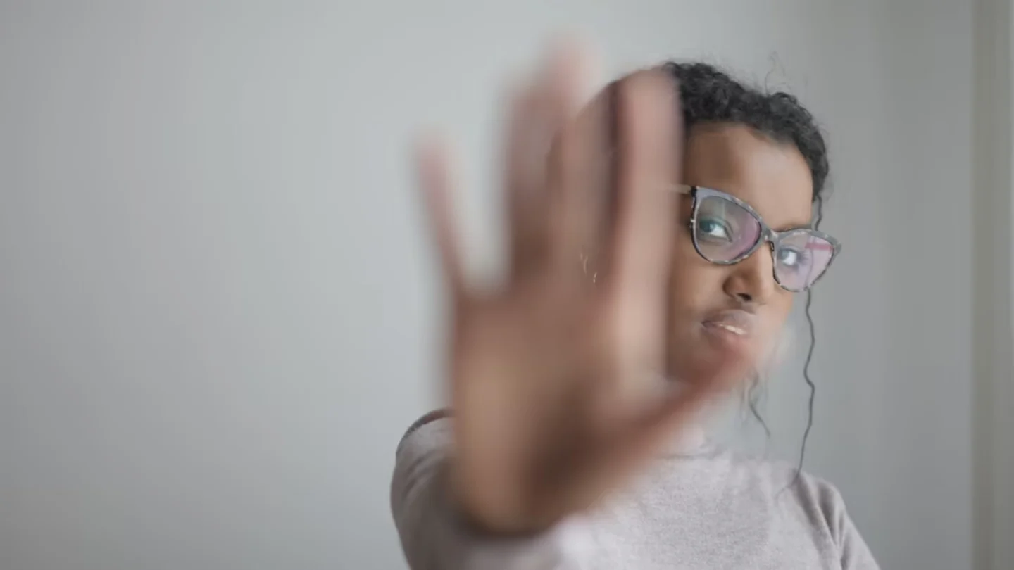 Young African American female wearing casual clothes and eyeglasses outstretching arm at camera while standing near grey wall