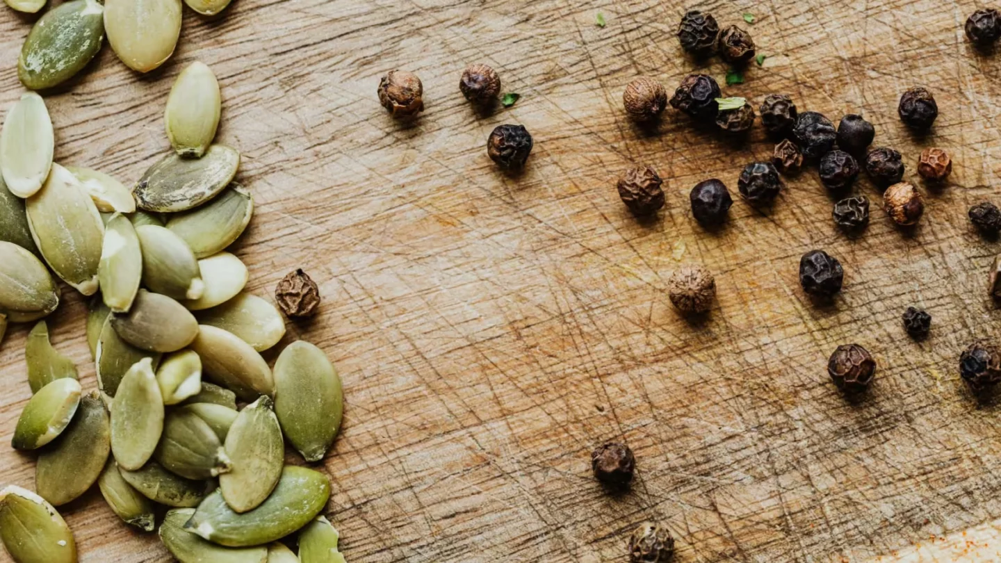 Close-up of pumpkin seeds and black peppercorns on a wooden cutting board.