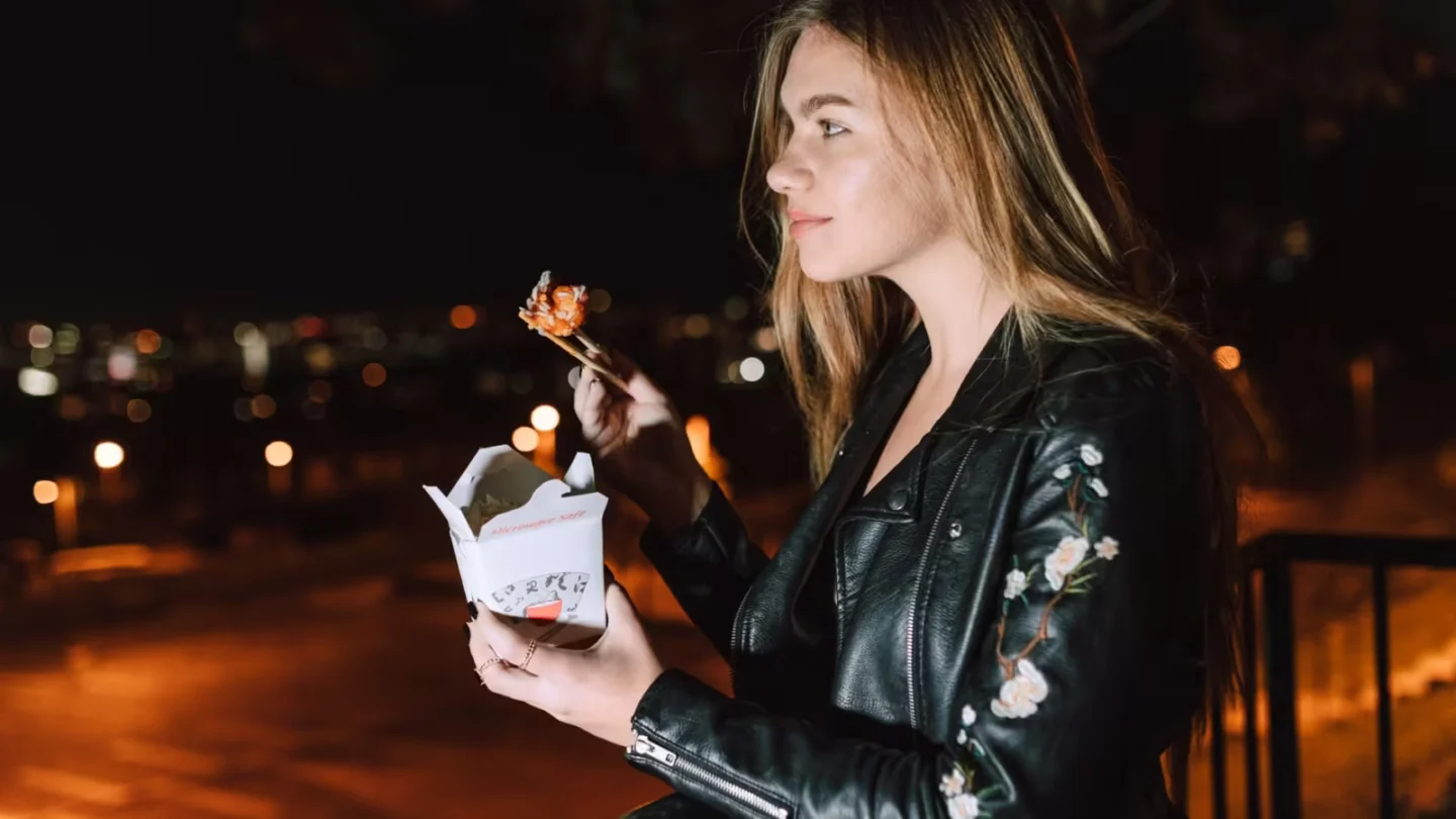 Young woman eating Asian takeout at night, city lights glowing in the background.