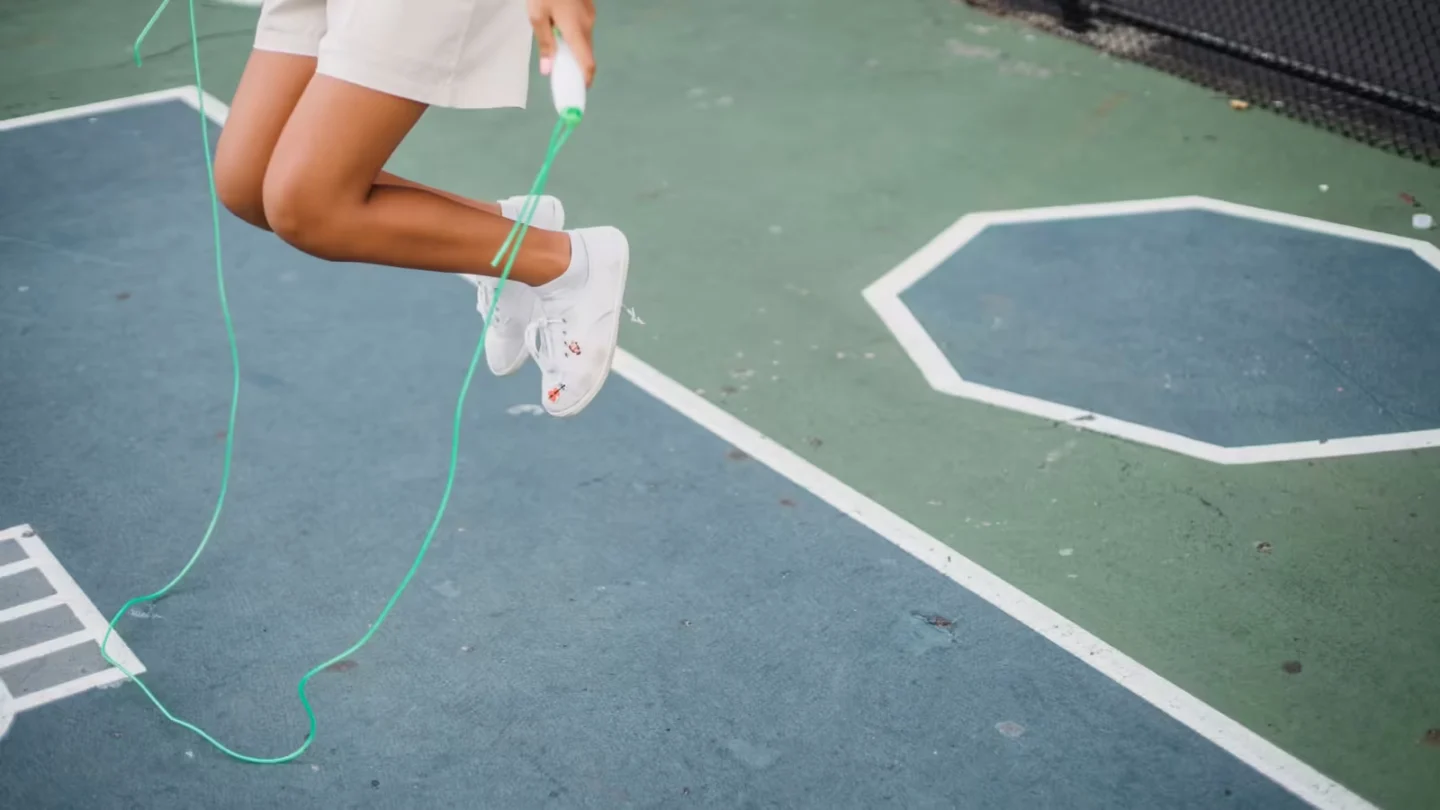 Girl playfully jumping rope on a painted outdoor playground surface, wearing casual shorts and sneakers.