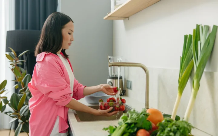 Asian woman washes strawberries at kitchen sink, promoting healthy living.
