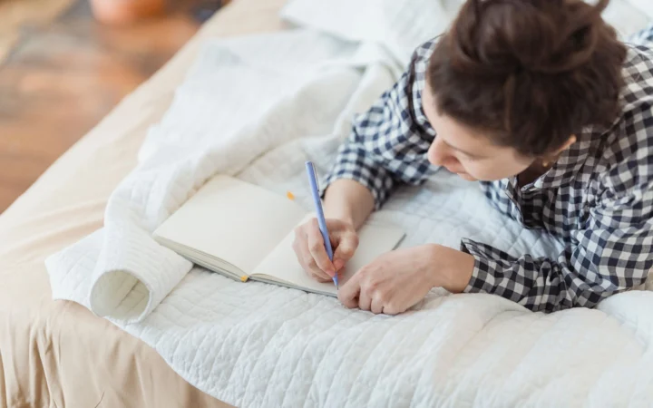 Woman writes in a journal while relaxing on a cozy bed with soft lighting and peaceful surroundings.