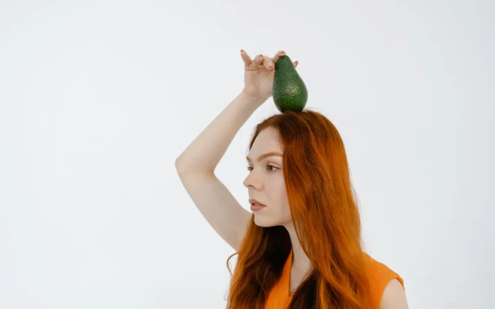 Woman with red hair balances an avocado on her head in a studio shot.