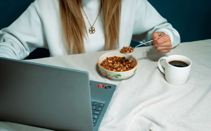 Woman enjoying cereal breakfast while browsing on laptop, featuring coffee and casual work setup.