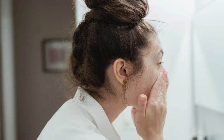 Young woman in a bathrobe follows her skincare routine, washing her face in a bathroom.