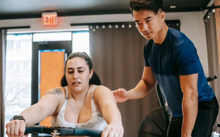 Young handsome ethnic experienced trainer supporting plus size woman working out on rowing machine in gym