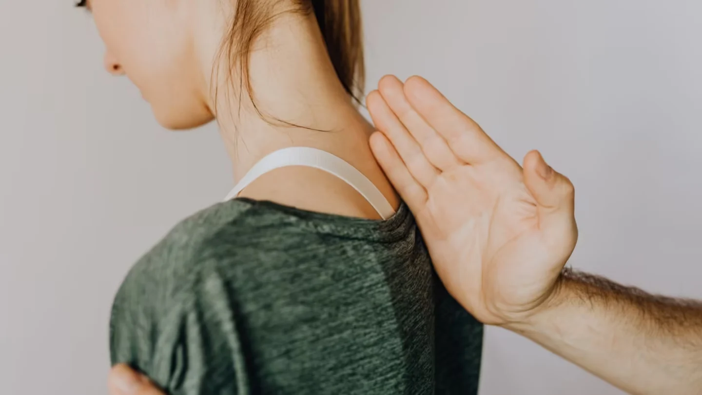 Side view of crop anonymous osteopath examining spine of slim young lady in casual wear near white wall in doctor office