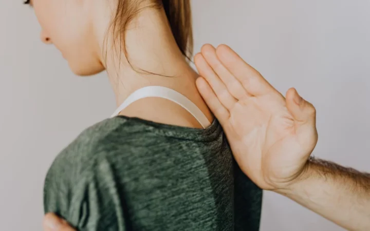 Side view of crop anonymous osteopath examining spine of slim young lady in casual wear near white wall in doctor office