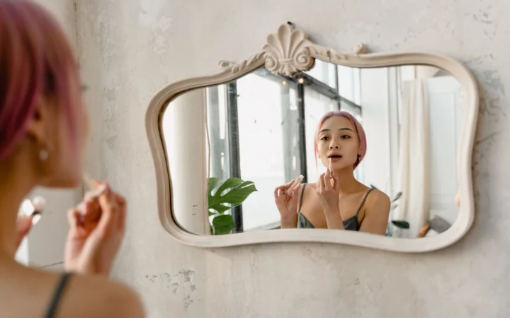 Young woman with pink hair applying makeup in front of ornate mirror indoors, embracing self-care.