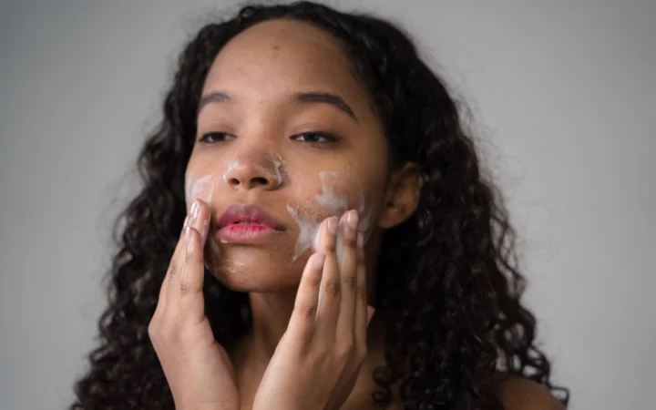 African American female with long dark curly hair washing cheeks with facial foam on gray background