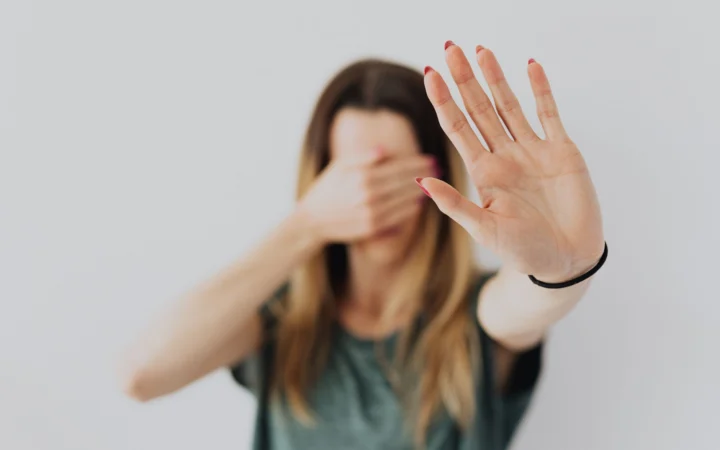 Woman in a gray shirt covers her face with her hand in a stop gesture, evoking a sense of fear and protection.