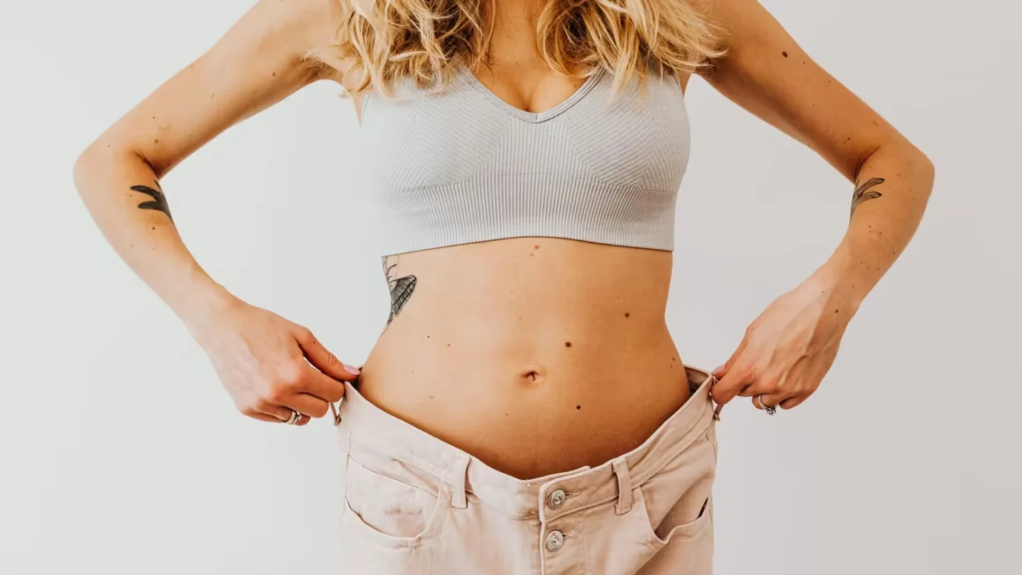 Woman displaying weight loss by wearing oversized pants and crop top, showing tattoos.