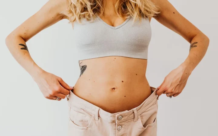 Woman displaying weight loss by wearing oversized pants and crop top, showing tattoos.