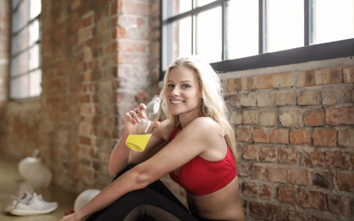 Young woman in workout attire smiling while holding a juice bottle indoors.
