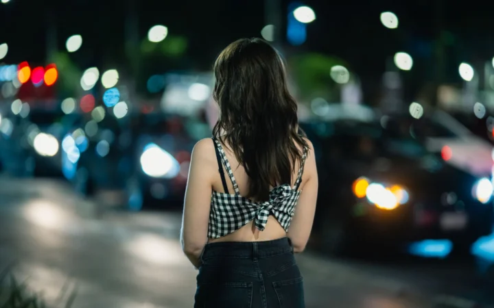 Woman with long hair stands in city street at night with blurred cars in background.
