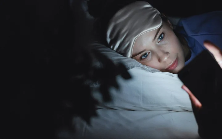 Woman in bed wearing a sleep mask uses her phone at night, creating a cozy ambiance.