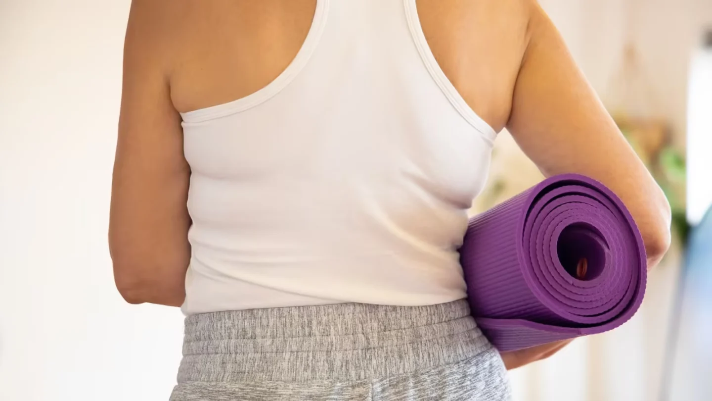 Woman in white tank top holding a rolled purple yoga mat indoors, preparing for exercise.