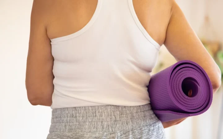 Woman in white tank top holding a rolled purple yoga mat indoors, preparing for exercise.