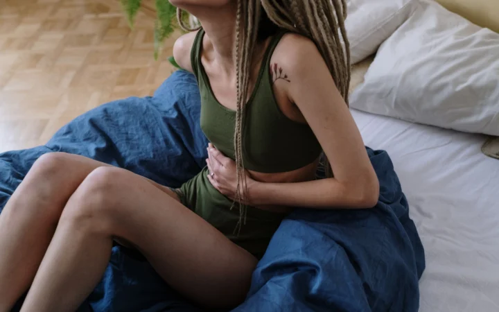 Woman sitting in bed holding her stomach, indicating pain or discomfort, possibly due to cramps.