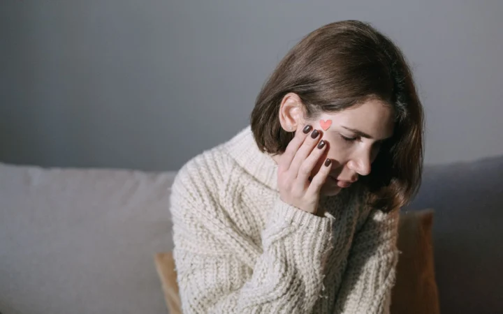 Woman wearing a sweater rests her hand on her face, displaying a heart sticker, in a softly lit room.