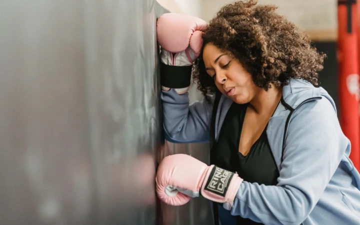 Side view of tired young African American female fighter with dark curly hair in sportswear and protective gloves leaning on