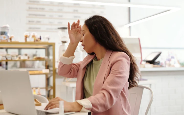 Young woman feeling unwell while working on her laptop indoors, looking fatigued.